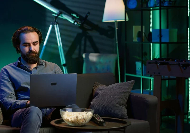 Homem assistindo filmes que todo amante de tecnologia deve assistir, com ambiente de tecnologia, iluminação de LED e um laptop no sofá.