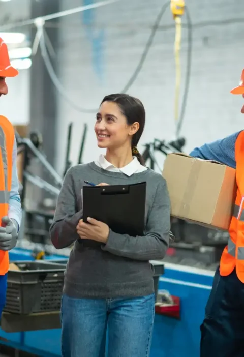 Equipe de trabalhadores em uma fábrica, incluindo duas pessoas de óculos de segurança e coletes de alta visibilidade, conversando e sorrindo, destacando a importância do bem-estar no trabalho para a segurança.