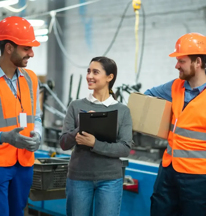 Equipe de trabalhadores em uma fábrica, incluindo duas pessoas de óculos de segurança e coletes de alta visibilidade, conversando e sorrindo, destacando a importância do bem-estar no trabalho para a segurança.