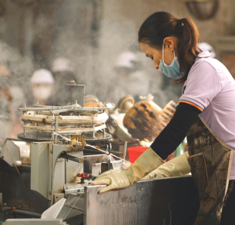 Trabalhadora com máscara, luvas e avental manuseia máquina industrial em área de produção com vapor ao redor.