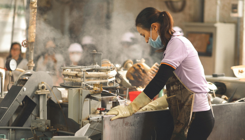 Trabalhadora com máscara, luvas e avental manuseia máquina industrial em área de produção com vapor ao redor.