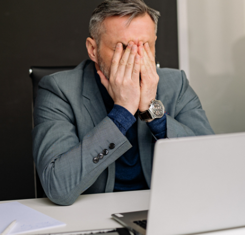 Homem de terno sentado à mesa cobre o rosto com as mãos em frente a um notebook, sugerindo estresse no trabalho.