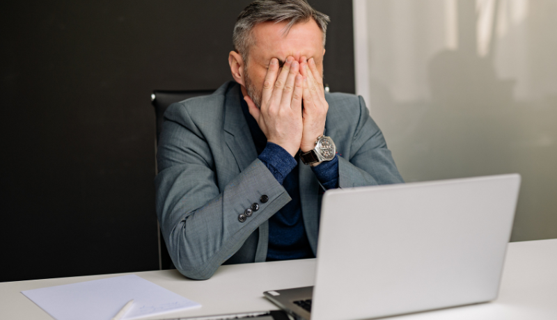 Homem de terno sentado à mesa cobre o rosto com as mãos em frente a um notebook, sugerindo estresse no trabalho.
