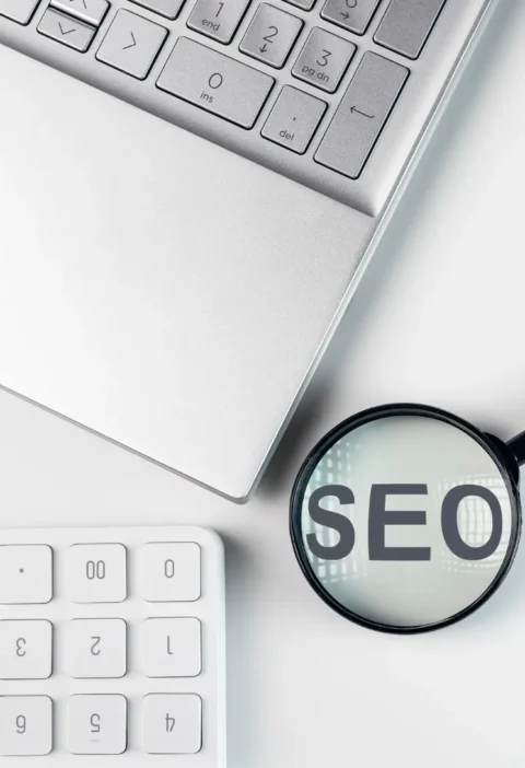 SEO programático: quando vale a pena gerar páginas em escala com templates e banco de dados — e quando isso vira spam: SEO em destaque dentro de uma lupa sobre um notebook e teclado, representando análise e otimização de buscas na internet.