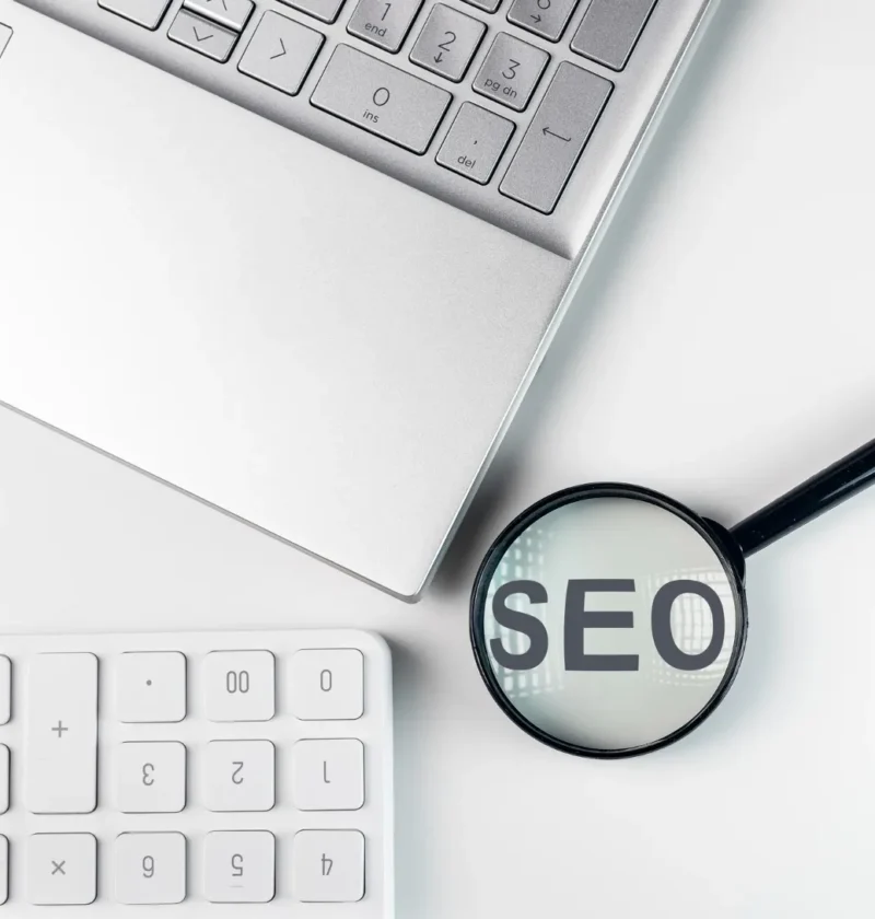 SEO programático: quando vale a pena gerar páginas em escala com templates e banco de dados — e quando isso vira spam: SEO em destaque dentro de uma lupa sobre um notebook e teclado, representando análise e otimização de buscas na internet.